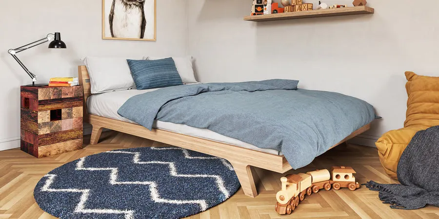 A walnut-framed platform bed with a blue duvet in a child's bedroom, with a nightstand, wall shelf, round rug, and wooden toy train nearby.