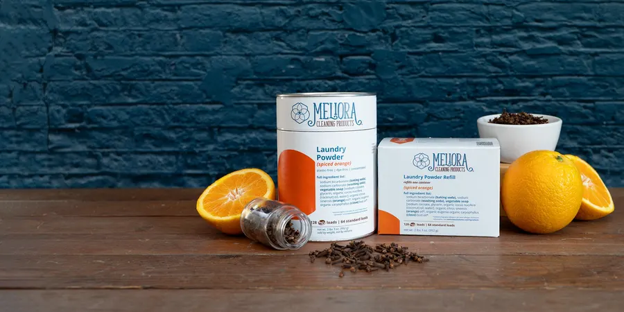 A canister and refill box of Meliora spiced-orange laundry powder on a wood table with fresh orange halves, whole oranges, and a small dish of whole cloves, against a dark blue brick wall.