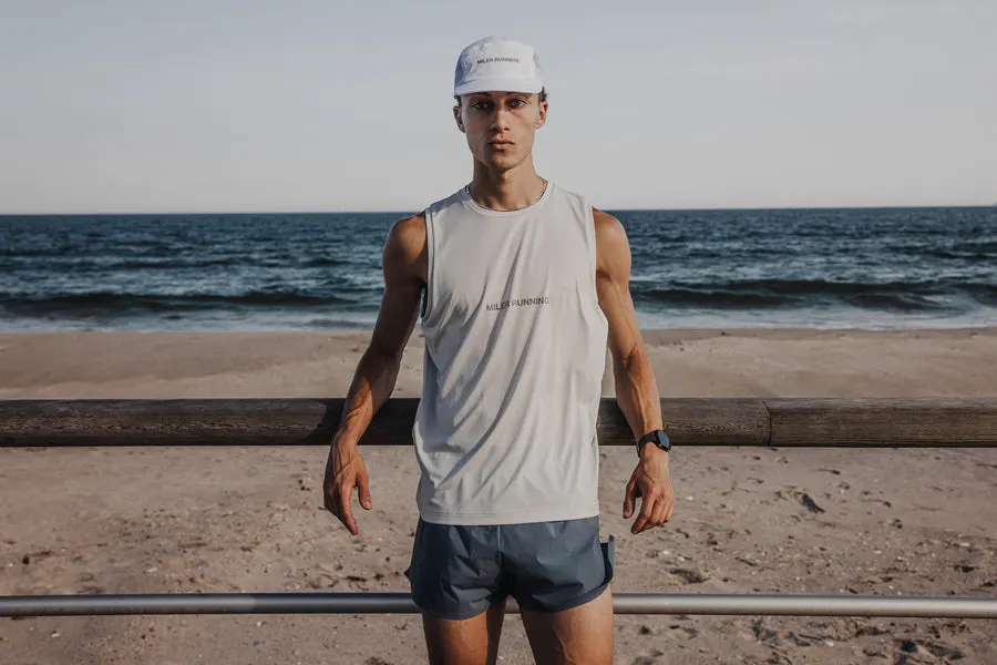 A young man in a light grey Miler Running tank top, running cap, and blue shorts stands on a beach boardwalk with the ocean behind him.
