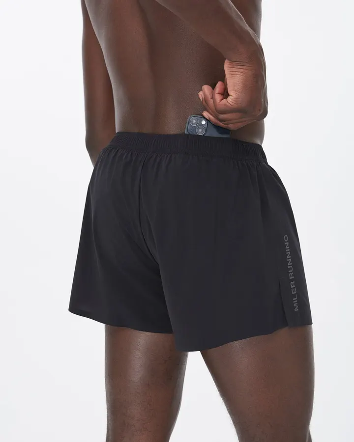 Close-up back view of a man wearing black running shorts with a reflective Miler Running side print, tucking a phone into the elastic waistband.
