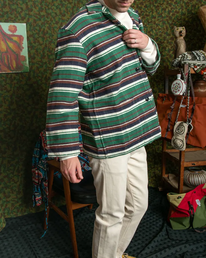 A man wearing a green, navy, cream, and rust striped flannel jacket over cream trousers, standing in a styled room with a camo-patterned wall, vintage bags, and folk-art objects on a shelf behind him.