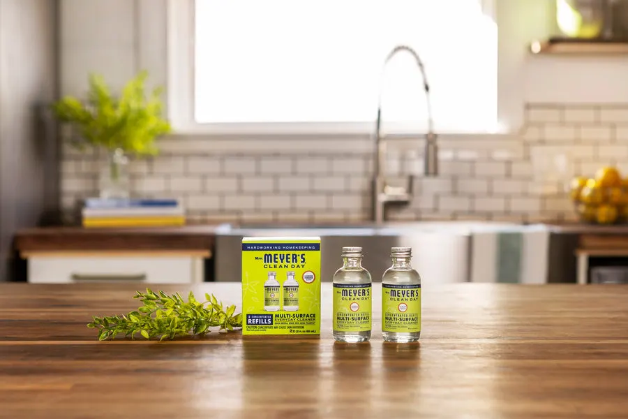 Two small glass bottles of Mrs. Meyer's Lemon Verbena Multi-Surface Everyday Cleaner concentrate refills and their yellow box, standing on a wood kitchen counter with a sprig of greenery, a tiled backsplash and sink blurred behind.