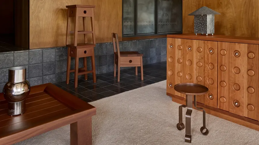 A wood-paneled showroom with a wooden sideboard, a stacked wooden stool, a chair, and a small round black metal side table arranged on a stone-tiled floor.