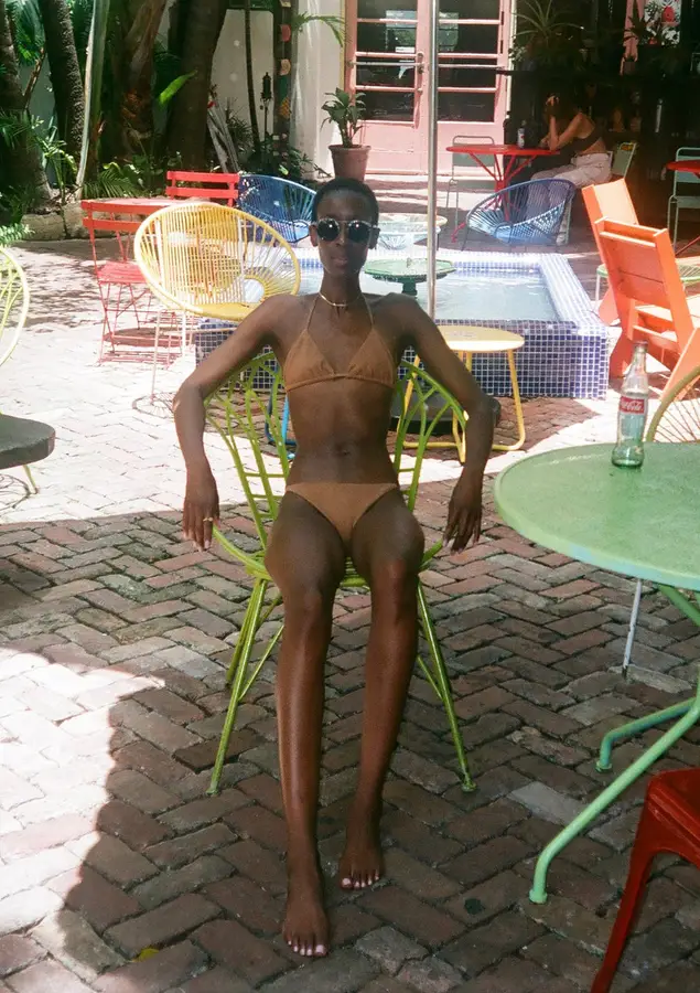 A woman in a tan triangle bikini sits on a green metal chair in a sunlit outdoor cafe courtyard with colorful chairs and a tiled fountain.