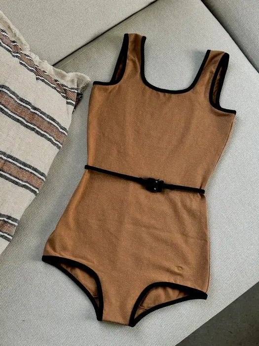 A tan one-piece swimsuit with black trim and a black belt buckle at the waist, laid flat on a grey cushion beside a striped blanket.