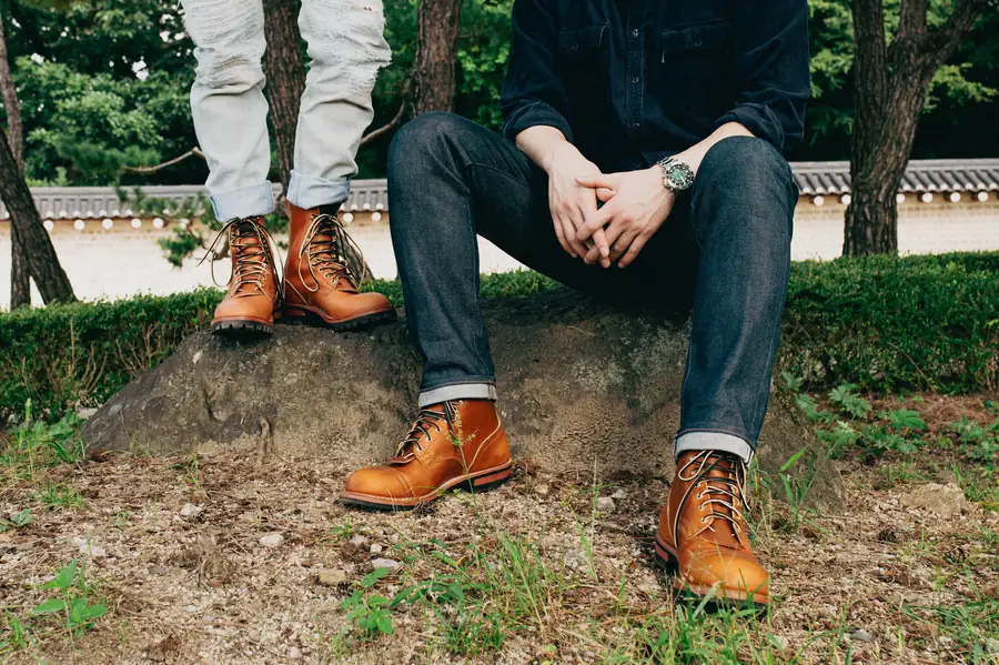 Two people on a mossy boulder in a park, cropped above the waist so only their legs show: both wear tan lace-up leather boots, one in cuffed raw denim and one in pale distressed jeans, with clipped hedges, pine trunks and a low white garden wall behind them.