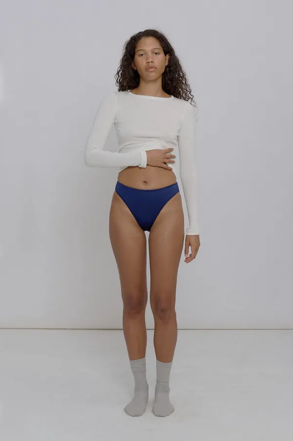 A woman with curly dark hair wears a navy high-cut bikini bottom with a white long-sleeve crop top, one hand resting on her hip, standing against a plain grey studio backdrop.