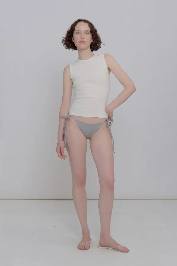 A woman in a cream ribbed sleeveless shell tank and grey tie-side bikini bottom, standing against a plain grey studio backdrop.