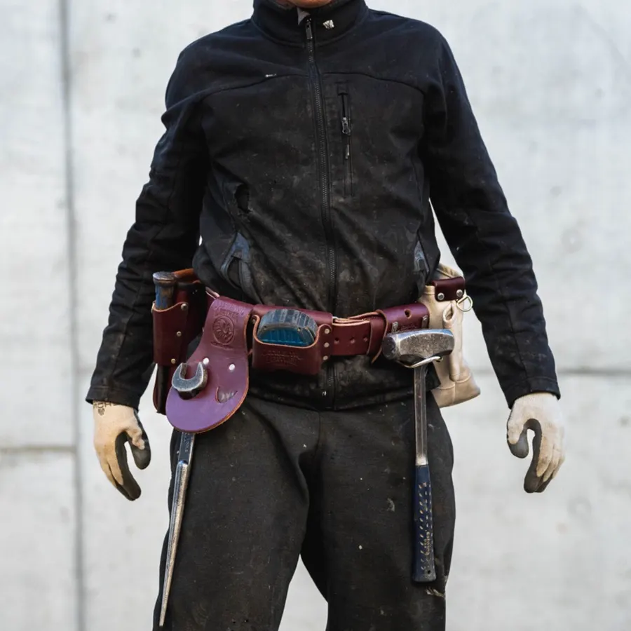 A worker in a black jacket and dirty work pants, face out of frame, wears a burgundy leather tool belt loaded with a wrench, hammer, and pouches, standing against a concrete wall.