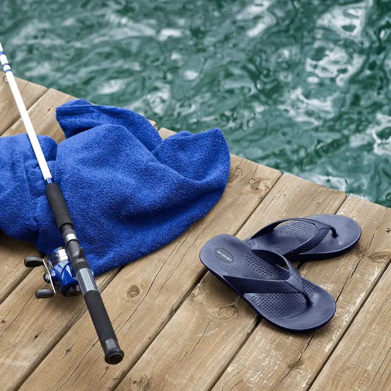 A pair of navy blue flip-flops on a wooden dock beside a rolled blue towel and a fishing rod and reel, with green water visible behind.