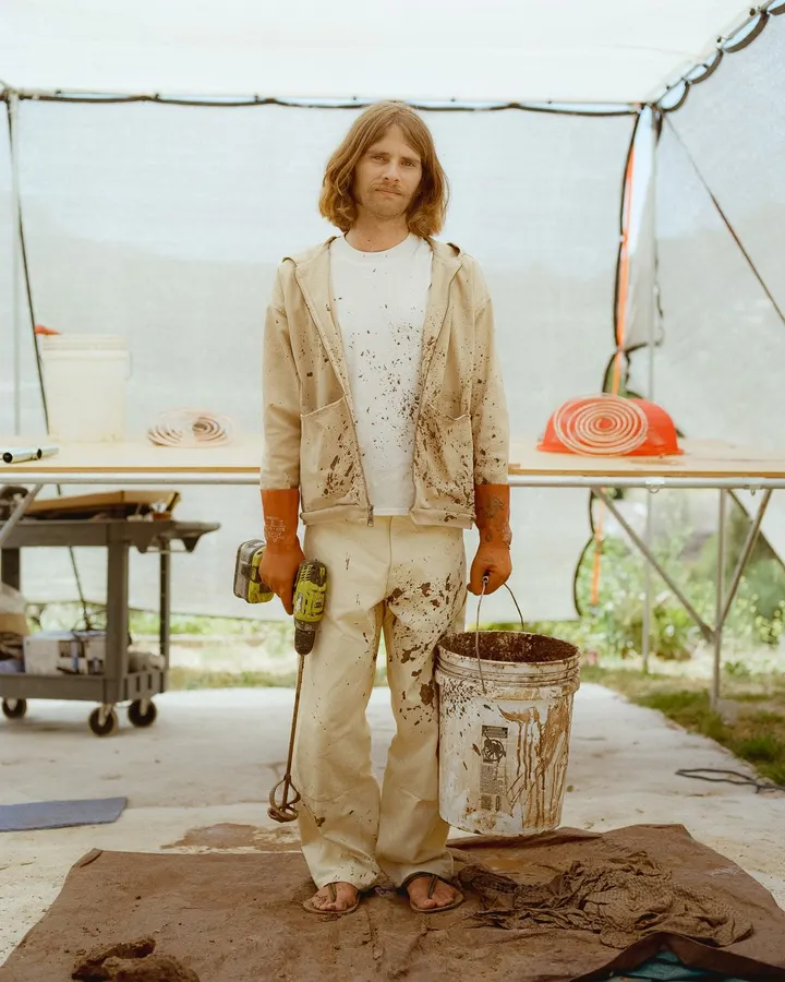 A long-haired man in mud-splattered cream workwear stands under an open-sided tent holding a power drill and a bucket of wet clay dye.