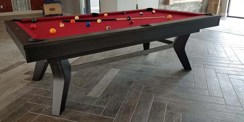 A dark wood pool table with red felt set up with balls racked mid-break in a modern living room with herringbone tile floors.