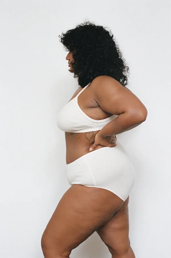 A woman with curly dark hair stands in profile wearing a cream bralette and high-waisted underwear against a plain light background.