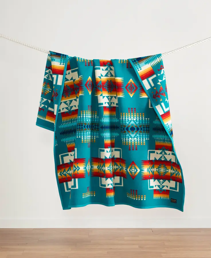 A turquoise wool blanket with a red, orange, and cream Chief Joseph geometric pattern, draped over a rope clothesline against a plain white wall above a wood floor.