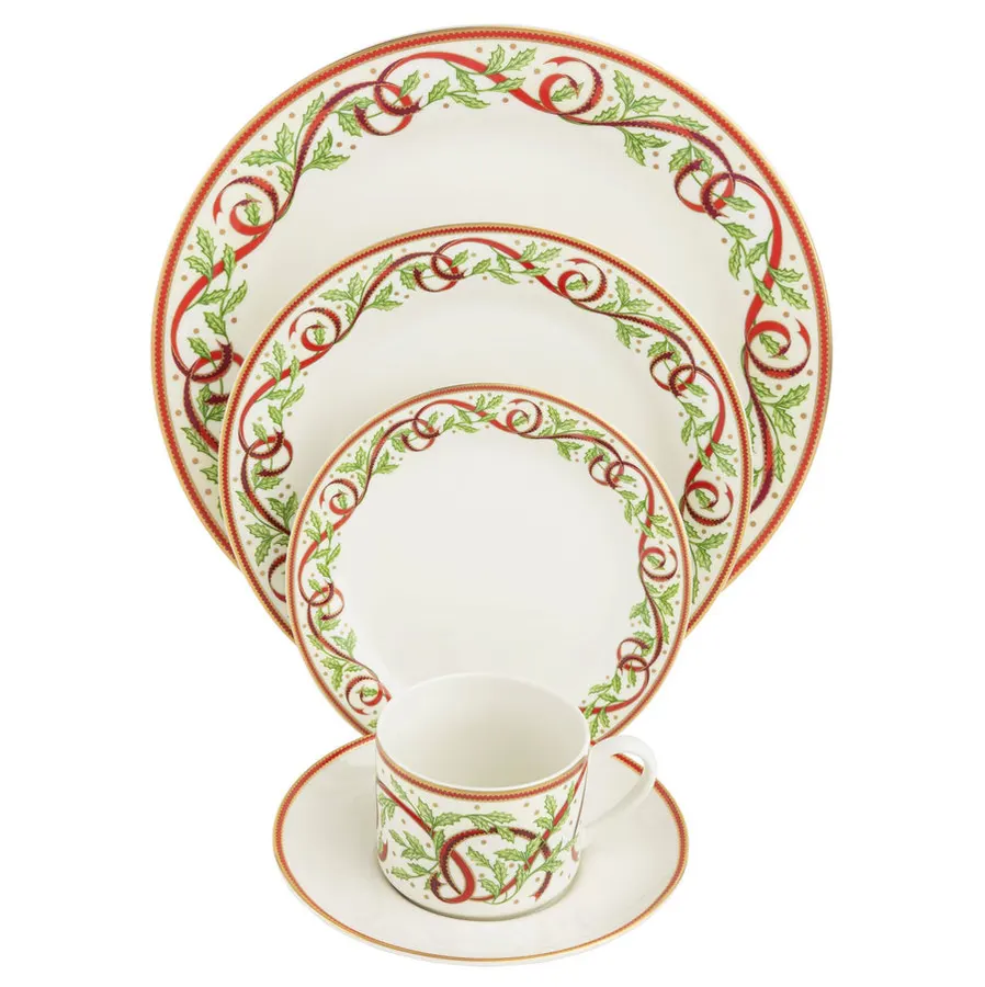 A five-piece white bone china place setting — dinner plate, salad plate, bread plate, teacup and saucer — decorated with a red ribbon and holly-leaf border and gold trim, fanned out and shot on white.