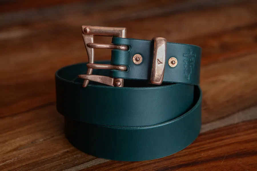 A coiled dark green leather belt with an antiqued brass roller buckle and copper rivets, resting on a wood-plank surface.