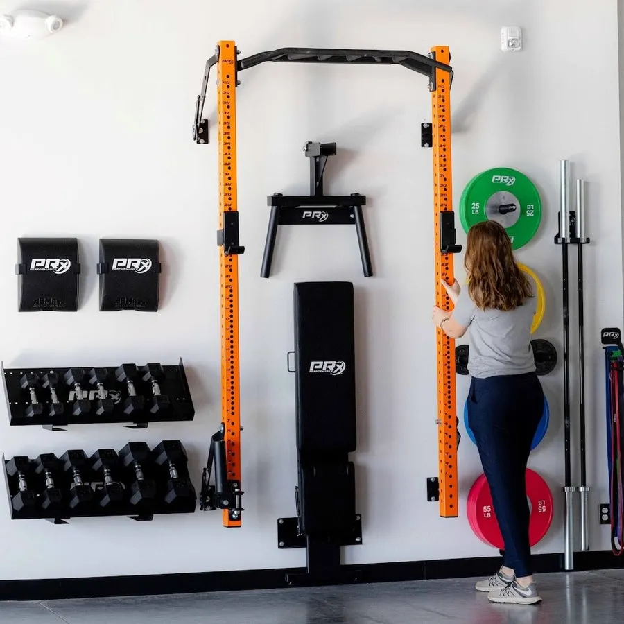 A woman in a home garage gym reaches to hang a weight plate on an orange wall-mounted folding squat rack, with dumbbells, resistance bands, and a barbell also mounted on the wall beside her.