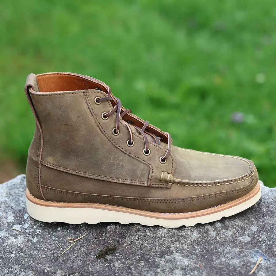 A single olive-brown suede moc-toe chukka boot with a cream lug sole, shown in profile resting on a lichen-covered rock with green grass blurred behind it.