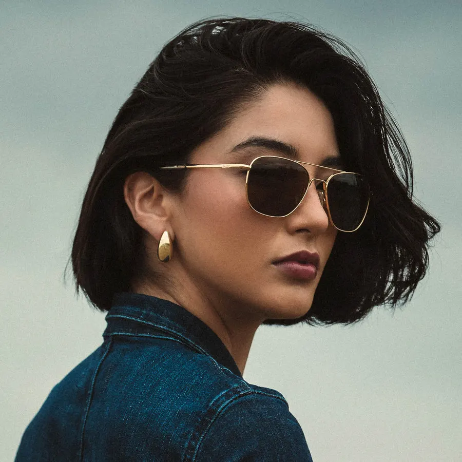 A woman with short dark hair looking over her shoulder outdoors, wearing gold-framed aviator sunglasses with dark lenses, a denim jacket collar visible.