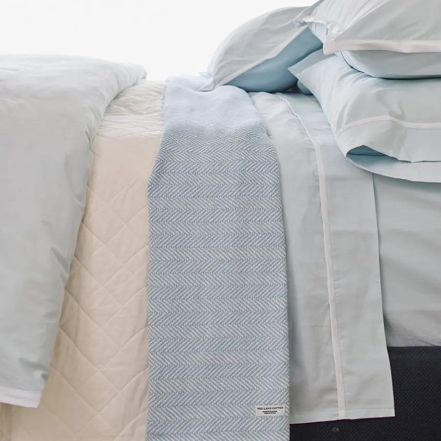 Pale blue cotton bedsheets and pillowcases layered on a made bed, with a folded herringbone-woven throw blanket carrying a small woven "Red Land Cotton" label draped across the foot.