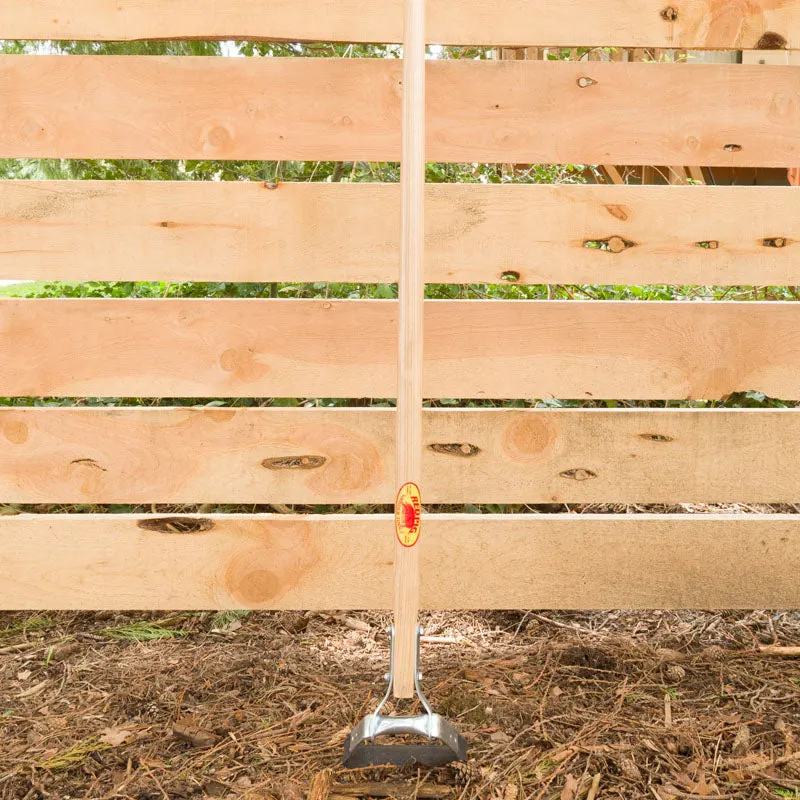 A long-handled steel stirrup hoe leaning upright against a cedar plank fence, its wooden handle stamped with a round red-and-yellow Red Pig maker's-mark sticker, set outdoors over mulch and greenery.