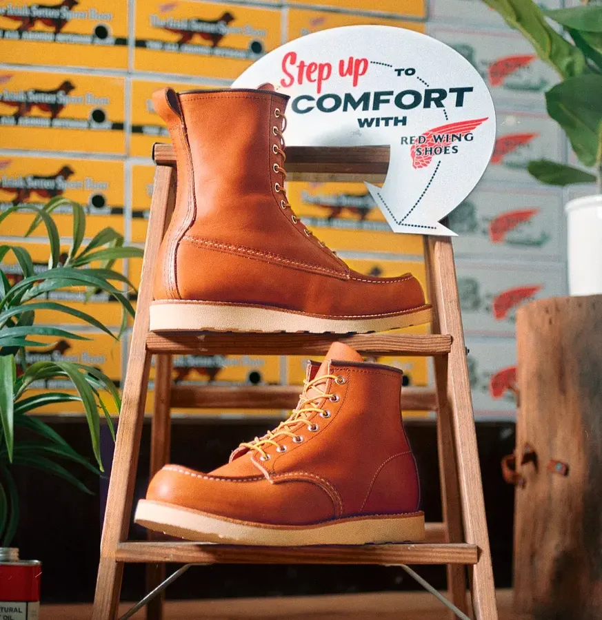 Two tan leather moc-toe work boots displayed on the rungs of a wooden step ladder, with vintage Red Wing Shoes advertising signage and potted plants in the background.