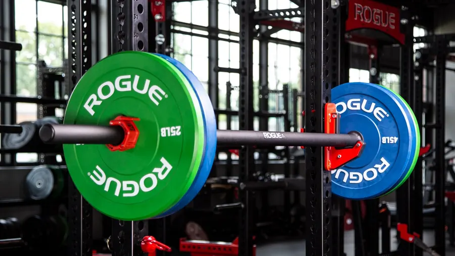 A black powder-coated Rogue axle bar racked in the J-cups of a steel rack, loaded at each end with a green 25 lb and a blue 45 lb Rogue bumper plate held on by red spring collars; more racks and a bank of windows sit out of focus behind it.