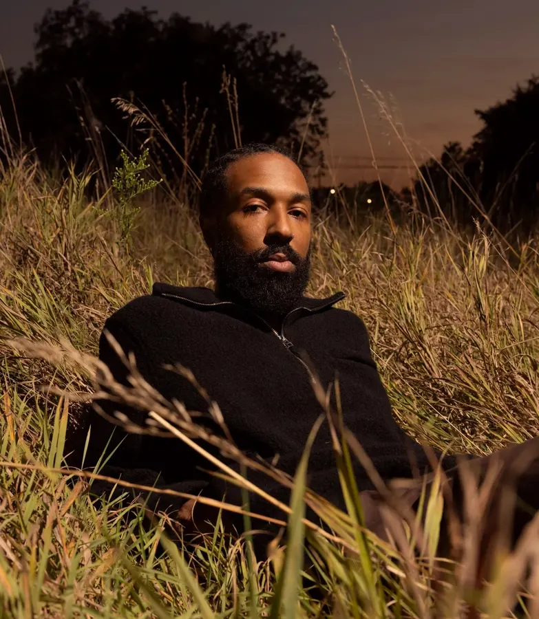 A man with a full beard reclines in tall dry grass at dusk, wearing a black quarter-zip fleece pullover, trees and a fading orange sky behind him.