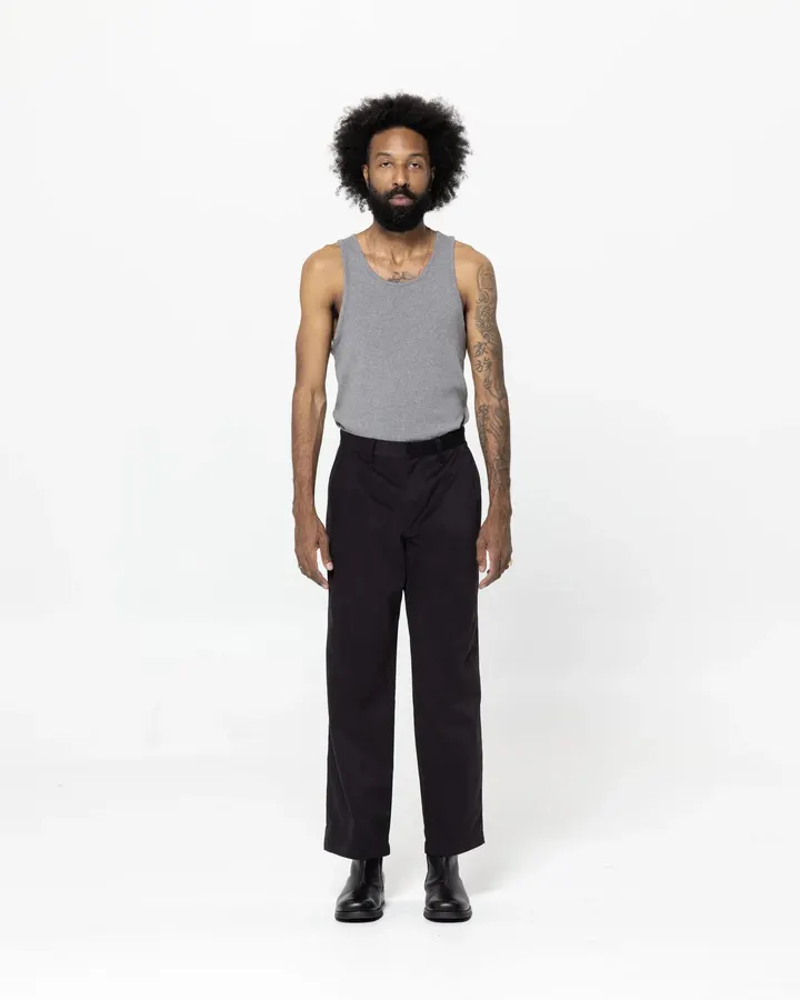 A man in a grey tank top and black straight-leg trousers with black Chelsea boots, standing against a white studio background.