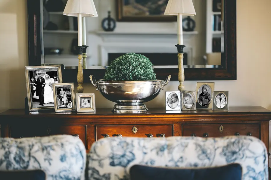A polished silver pewter bowl holding a small green topiary, flanked by candlesticks and framed family photographs, on a wooden sideboard beneath a mirror.
