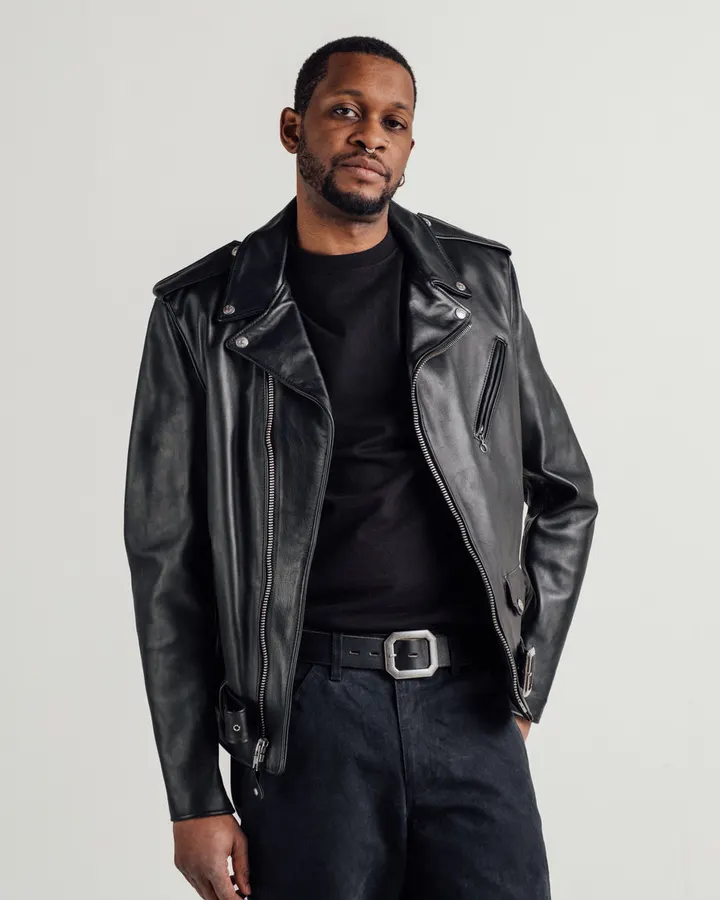 A man with a beard wearing an open black leather motorcycle jacket over a black t-shirt and dark trousers with a large silver belt buckle, shot against a plain studio background.