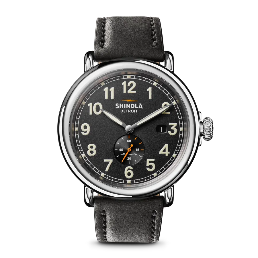 A Shinola Runwell automatic watch with a black dial, cream numerals, an orange seconds hand on a small sub-dial, and a black leather strap, photographed face-on against a white background.
