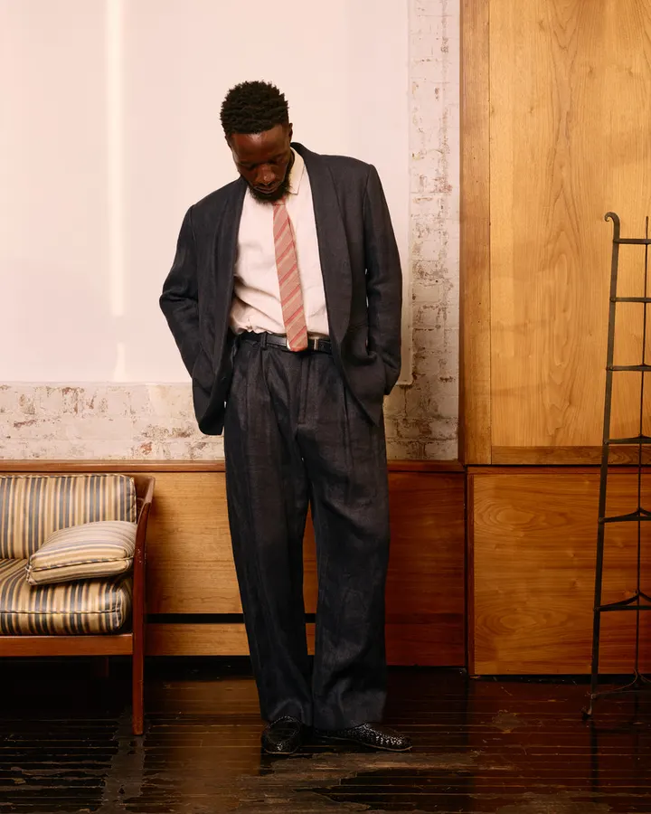 A man in a dark tailored suit jacket, white shirt, and striped tie stands with hands in pockets looking down, in front of a striped vintage sofa and wood-paneled wall.