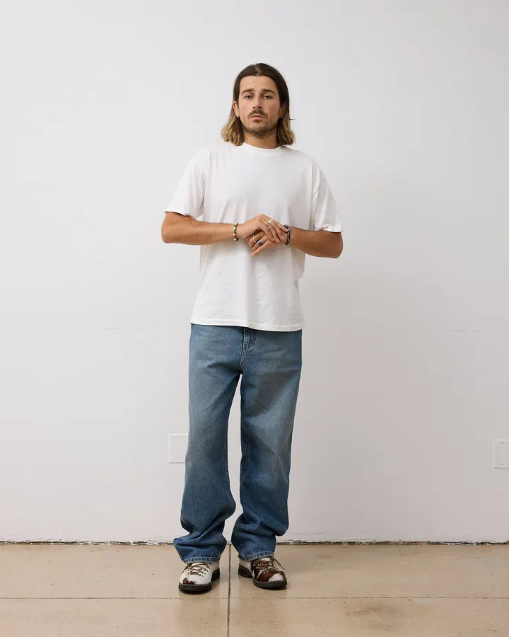 A man with shoulder-length hair wearing a plain white crewneck t-shirt and blue jeans, standing against a white wall.