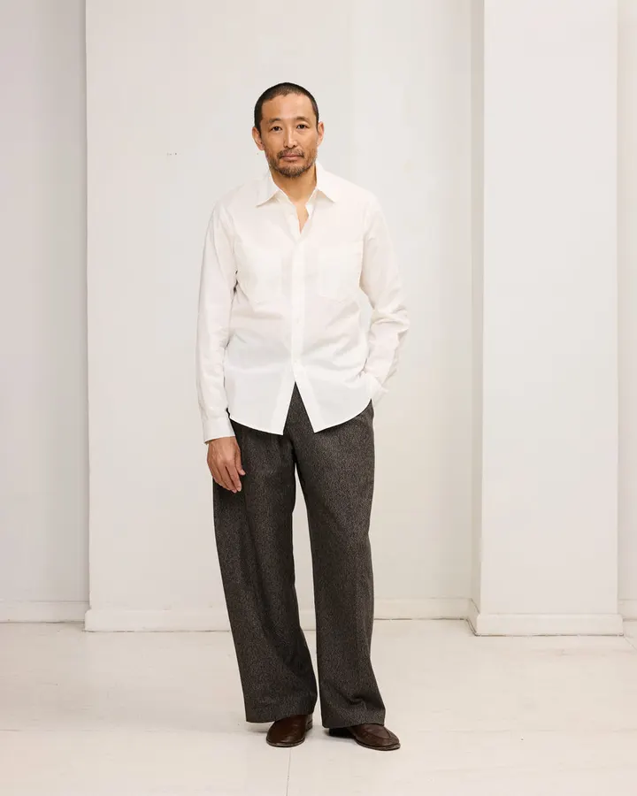 A man wearing an off-white button-down dress shirt with a Western-style yoke and grey wide-leg trousers, standing in a bright studio hallway.