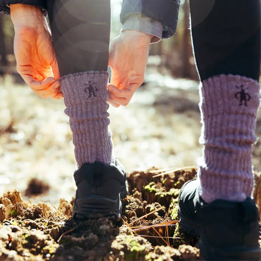 Two hikers bent over on a sunlit dirt trail, each pulling up a lavender ribbed wool hiking sock with the Smartwool logo over black hiking boots.