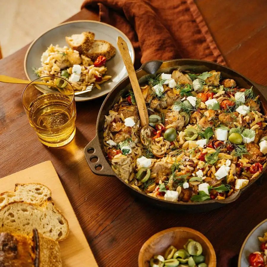 A Smithey No. 12 cast iron skillet filled with a chicken, orzo, olive, feta, and cherry tomato dish on a rustic wood table, beside a plate of the same food, a glass of white wine, torn bread on a board, and a bowl of olives.