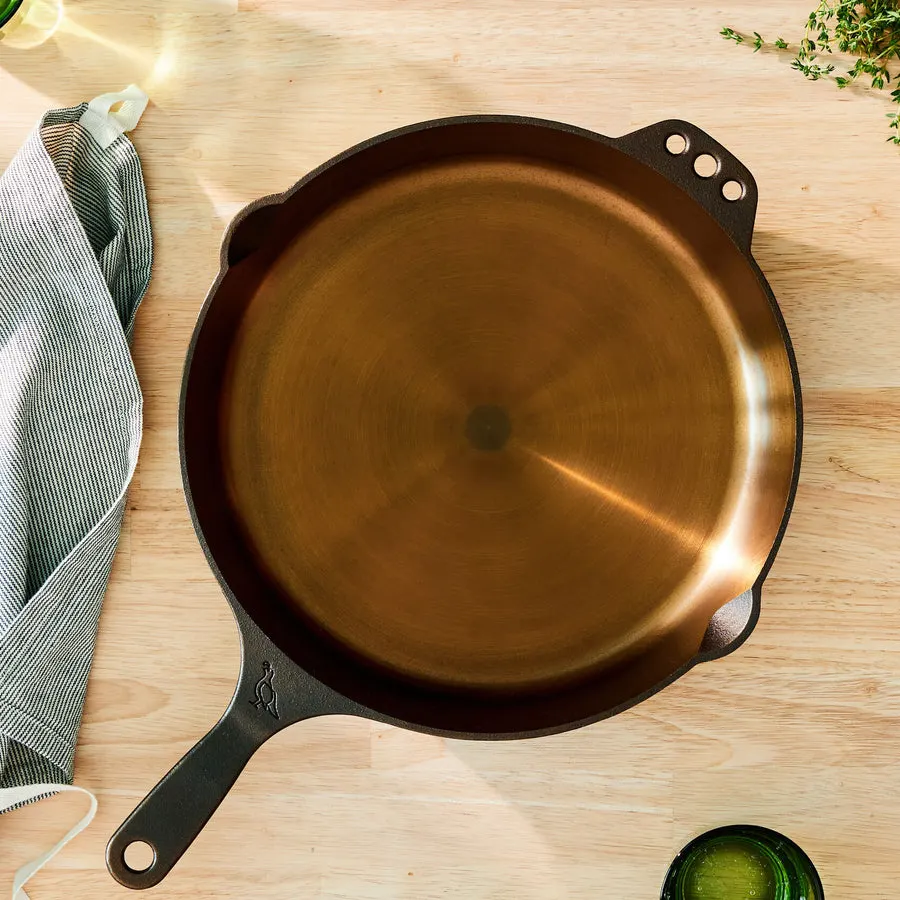 An empty, well-seasoned Smithey No. 12 cast iron skillet with a polished bronze-toned interior, viewed from above on a light wood cutting board next to a striped kitchen towel.