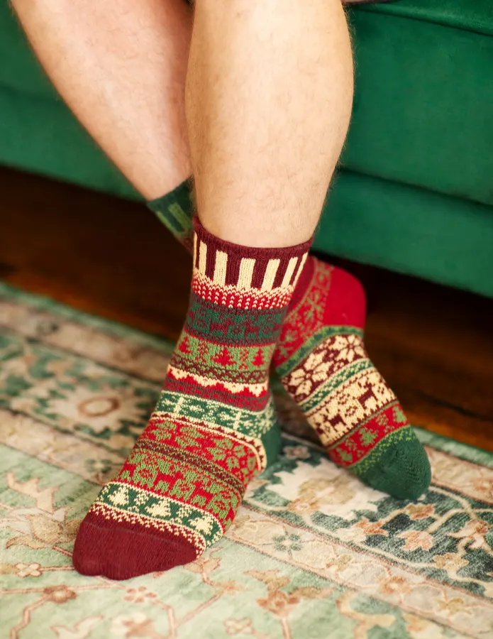 A pair of crossed legs wearing mismatched red, green and cream fair-isle patterned crew socks, feet resting on a patterned rug in front of a green upholstered seat.