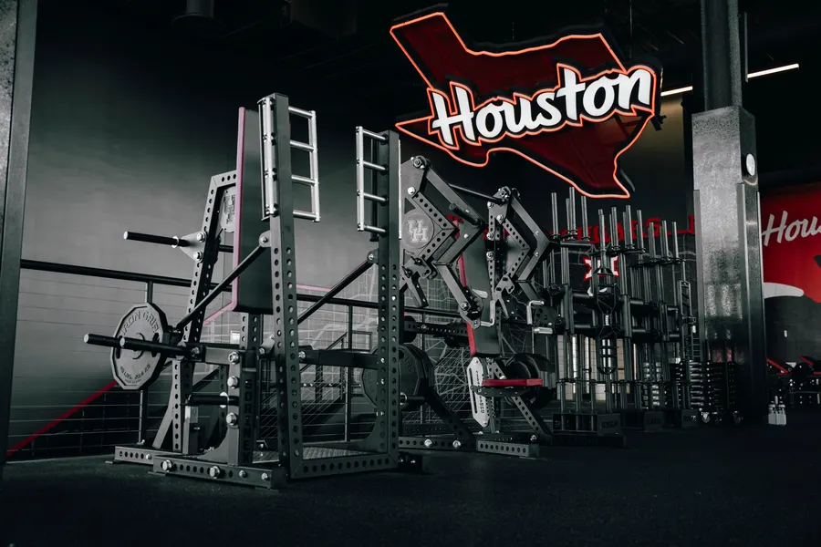 A row of black steel Sorinex power racks and specialty attachments in a dramatically lit University of Houston strength and conditioning facility, with a red-and-white Houston wordmark logo on the wall behind.