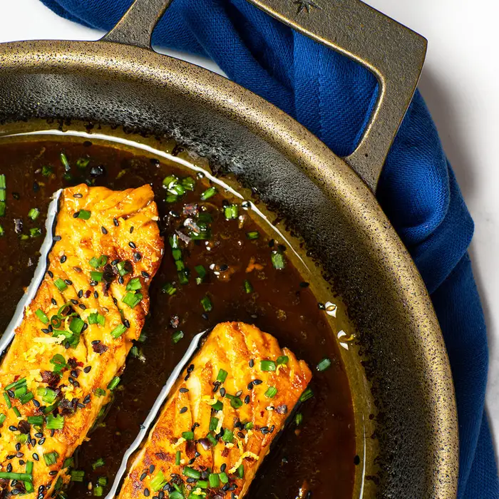 Overhead close-up of two glazed teriyaki salmon fillets topped with sesame seeds and chives, simmering in dark sauce in a bronze-finished cast iron skillet with the Stargazer star logo on its handle, resting on a blue linen napkin.