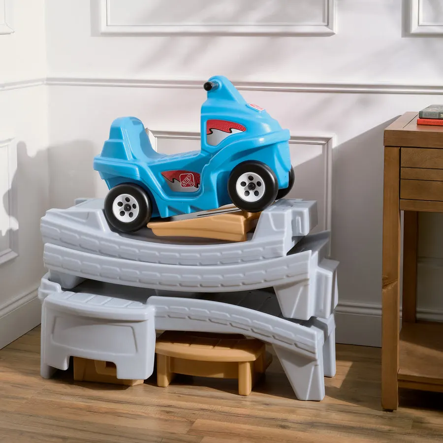 A blue plastic ride-on toy car sits atop a partly folded grey roller-coaster track set staged in the corner of a sunlit room, next to a wooden side table.