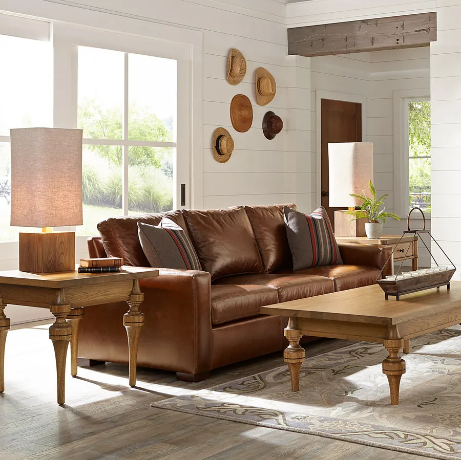 A sunlit farmhouse-style living room with a cognac-brown leather Stickley sofa, striped throw pillows, oak side tables and coffee table, a patterned area rug, and straw hats hung on a shiplap wall.