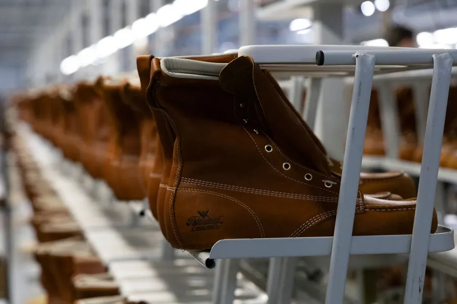 Rows of tan leather Thorogood work boots hang from a white conveyor rack on a factory production line, with more boots blurred in the background under overhead lights.