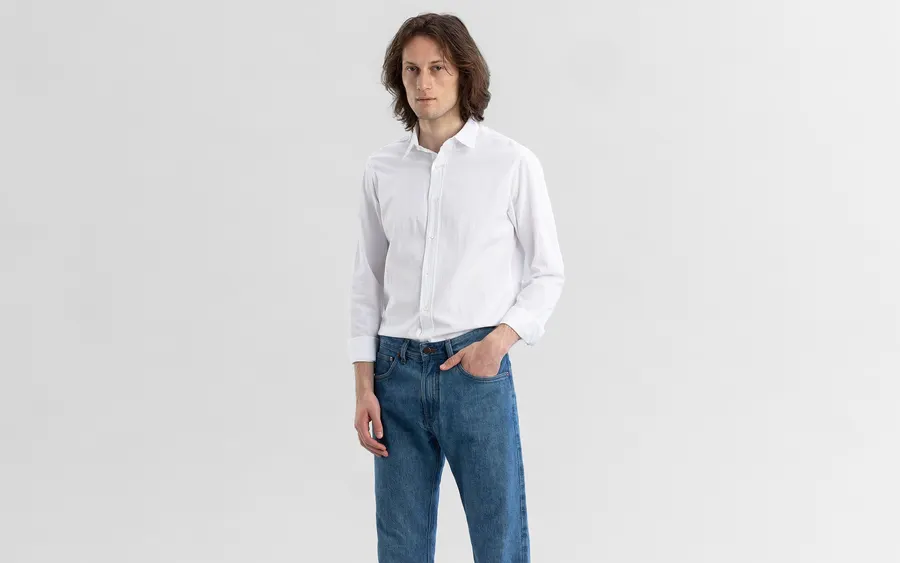 A man with shoulder-length brown hair stands against a plain light grey studio backdrop, wearing a white long-sleeve button-up shirt tucked into medium-wash blue jeans, one hand in his pocket.