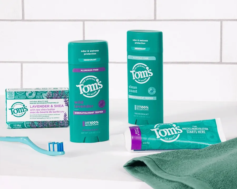 A bathroom counter with a bar of Tom's of Maine lavender and shea soap, two deodorant sticks, and a toothpaste tube resting on a green washcloth, with a blue toothbrush in front, against white subway tile.