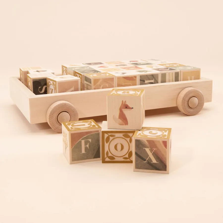 A wooden toy wagon loaded with illustrated alphabet blocks, with several loose blocks including a fox-print block spelling F-O-X stacked in front on a pale pink background.