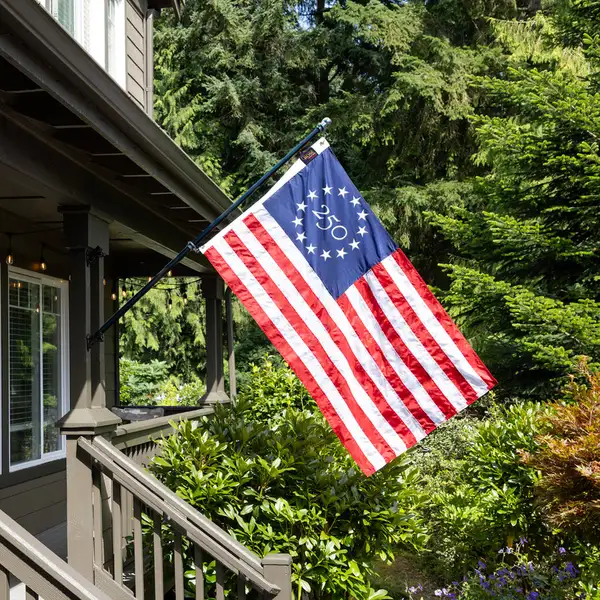 An embroidered American flag with a circular 250 emblem in the canton flies from a porch-mounted pole in front of a house, framed by evergreen trees and garden shrubs.