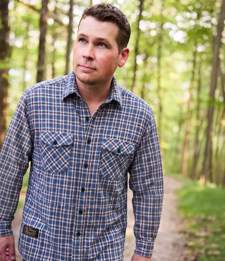 A man in a blue, brown and cream plaid flannel shirt looking off to the side while walking on a wooded trail with dappled sunlight through the trees.