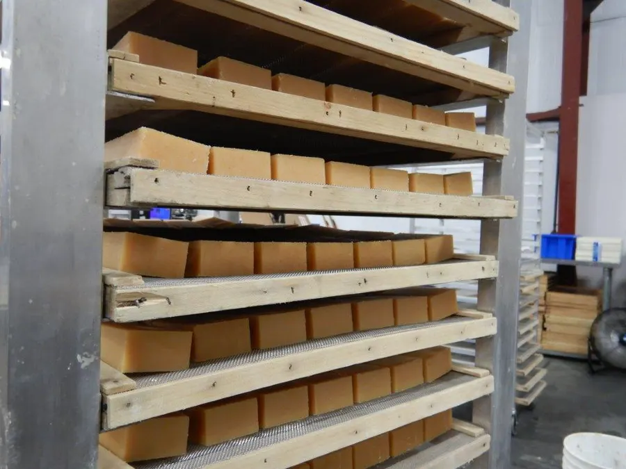 Rows of tan bar soap resting on wooden slatted curing racks in a warehouse, seen from a low angle looking down the length of the rack.